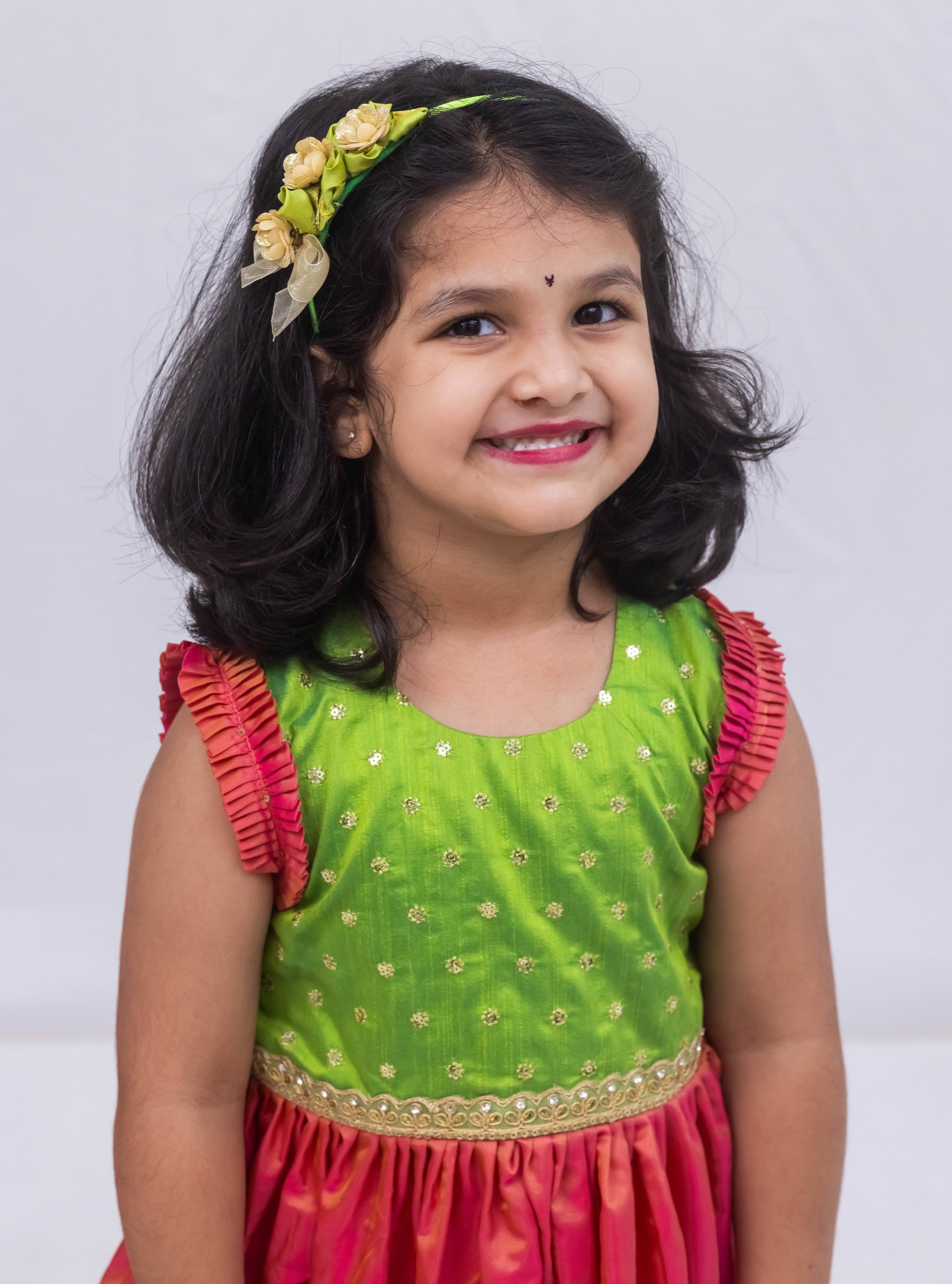 This exquisite frock for girls features a Parrot Green sequined bodice adorned with delicate lace detailing, paired with a luxurious Blush Pink Mysore silk skirt. The armholes are accentuated by intricately pleated frills that are adorned with matching sequins for a stunning finish.