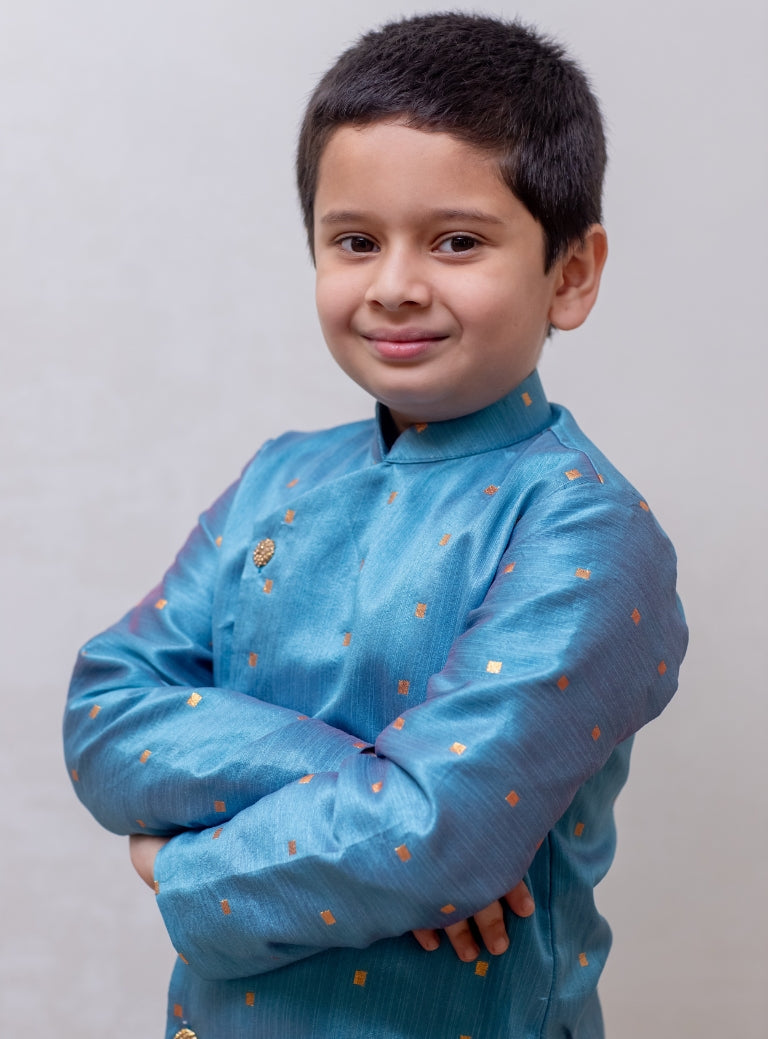 This boys' kurta features a Steel Blue mulberry silk fabric with a front open stand collar and distinctive stone-studded buttons. It is paired with a Golden Banglori silk salwar, and the kurta includes a comfortable cotton lining for added comfort.