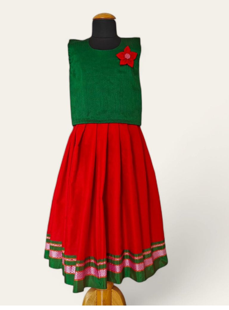 This creation features a handloom red khunn parkar with a striking green border, paired with a green raw silk sleeveless blouse. The blouse is adorned with handcrafted flower motifs, making it a perfect choice for girls.
