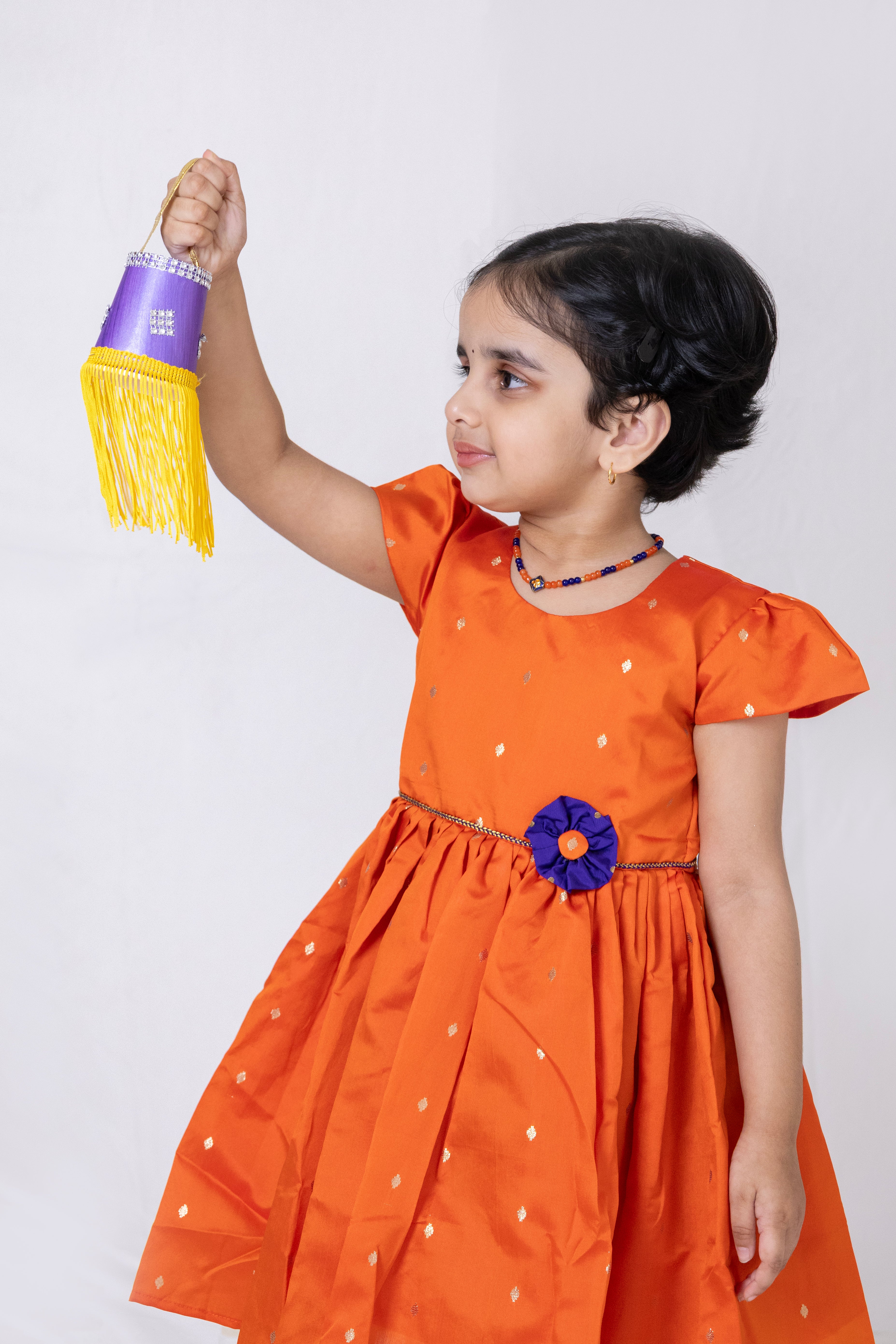 Orange taffeta silk flared dress with handmade purple fabric flower motif and chord waist belt for Girls.Let your princess be as comfortable as in her casuals with carefully designed and crafted Comfort Ethnic Wear by Soyara Ethnics. Keep her fashion quotient high with timeless patterns, vibrant combinations and royal textiles.