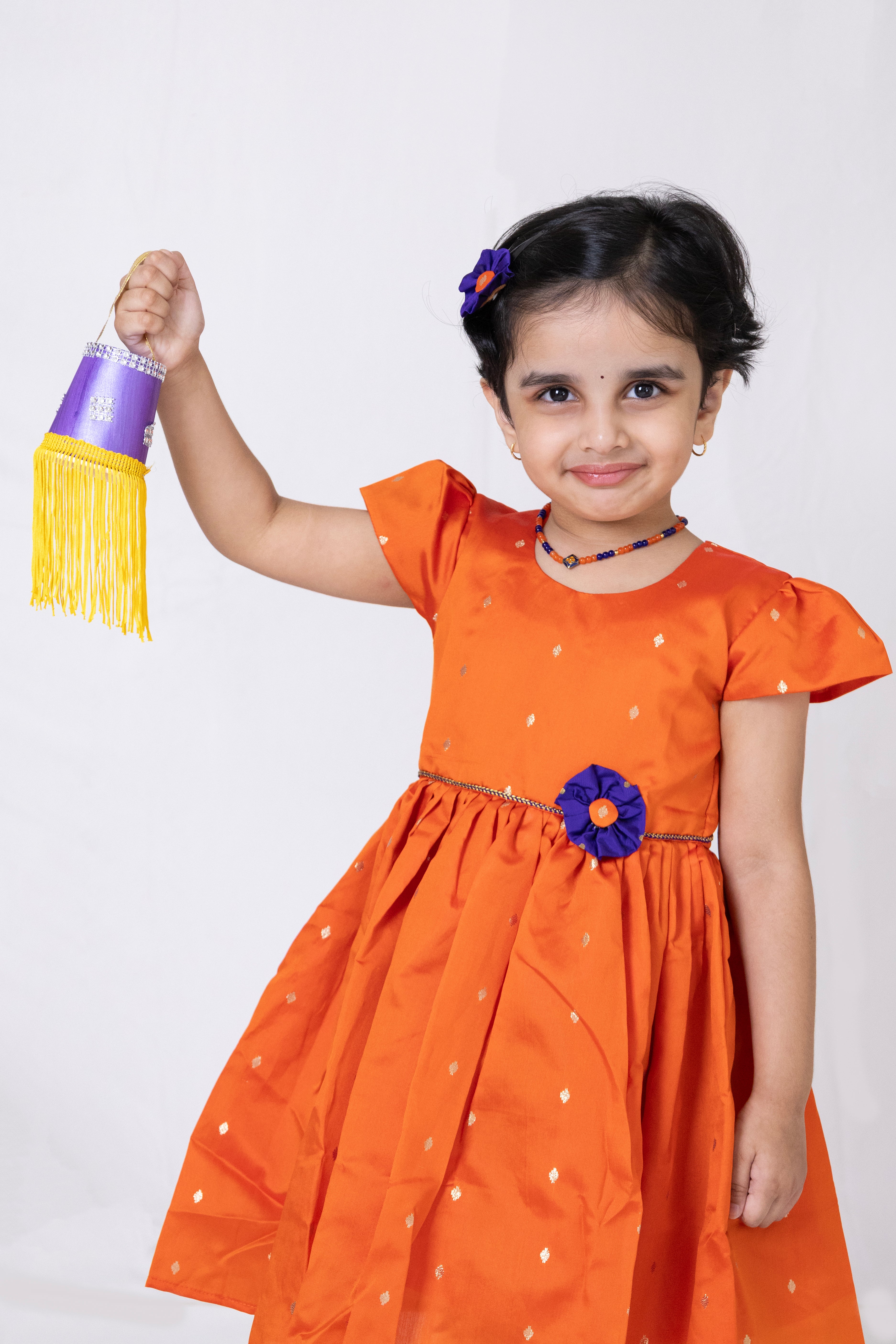 Orange taffeta silk flared dress with handmade purple fabric flower motif and chord waist belt for Girls.Let your princess be as comfortable as in her casuals with carefully designed and crafted Comfort Ethnic Wear by Soyara Ethnics. Keep her fashion quotient high with timeless patterns, vibrant combinations and royal textiles.