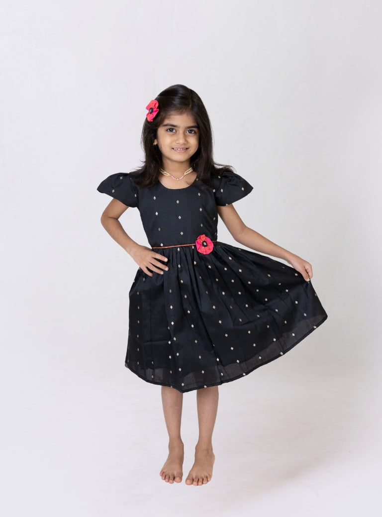 This girls’ flared dress features vibrant black taffeta silk, adorned with a handcrafted pink fabric flower motif and a chord waist belt. It is designed with elegant puffed cap sleeves for a refined finish.