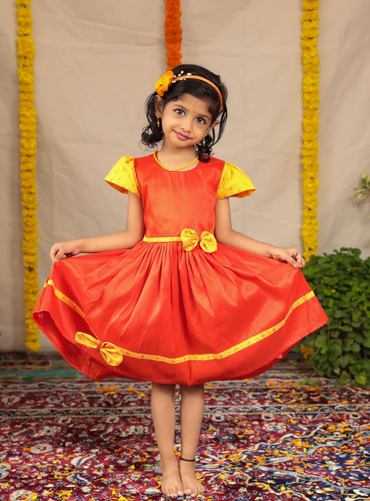 Orange Raw silk dress with yellow brocade sleeves and bow highlights for girl.Let your princess be as comfortable as in her casuals with carefully designed & crafted Comfort Ethnic Wear by Soyara Ethnics.Keep her fashion quotient high with timeless patterns, vibrant combinations and royal textiles.