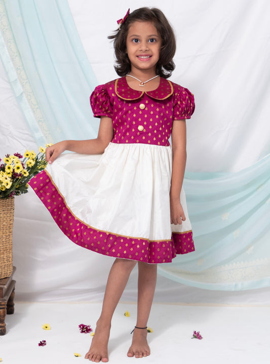This dress features a combination of magenta brocade silk and ivory Mysore silk, complete with a Peter pan collar and puffed sleeves. The bodice is adorned with contrasting buttons for a touch of elegance.