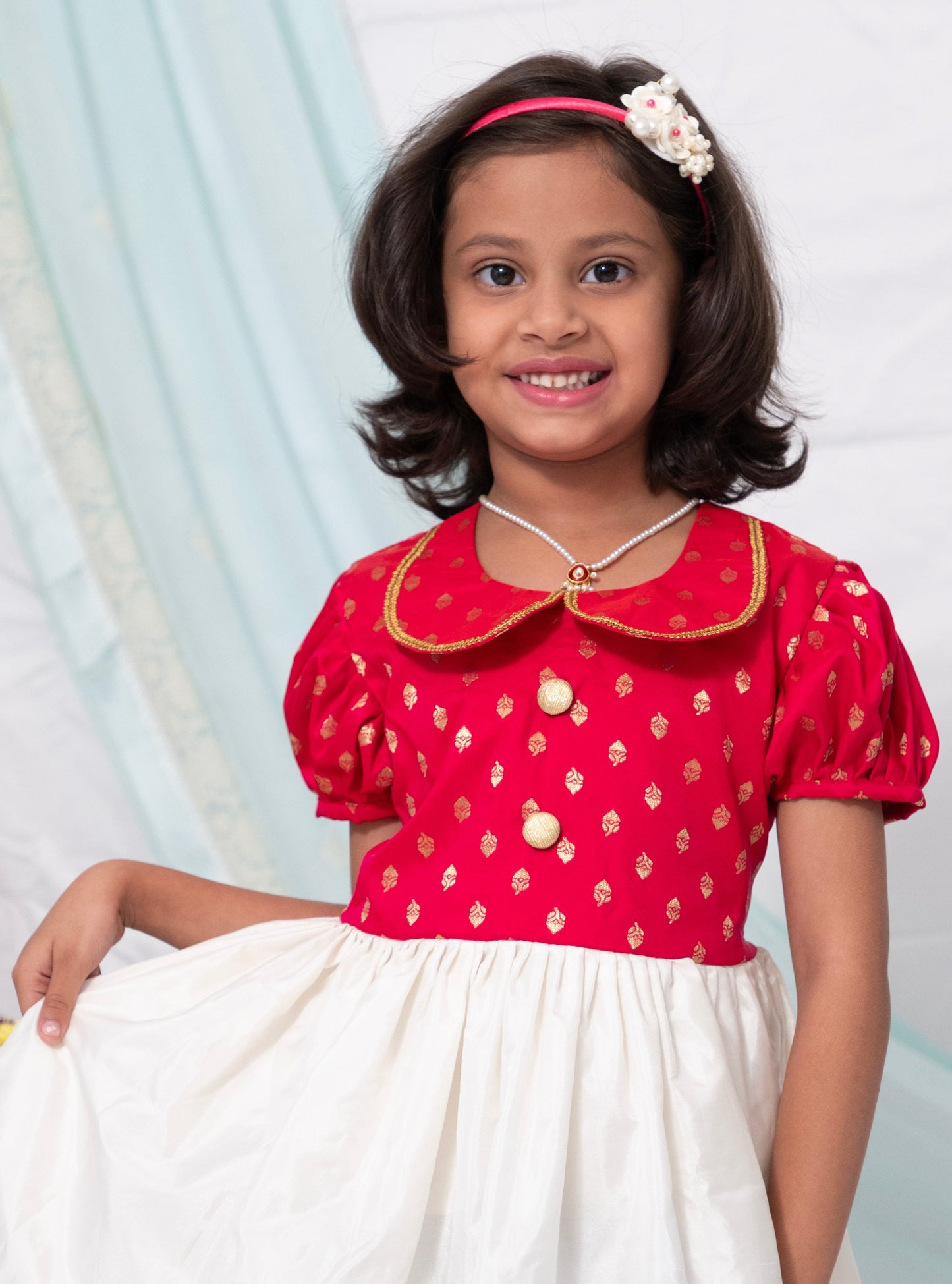 This dress features a combination of fuschia brocade silk and ivory Mysore silk, complete with a Peter pan collar and puffed sleeves. The bodice is adorned with contrasting buttons for a touch of elegance.