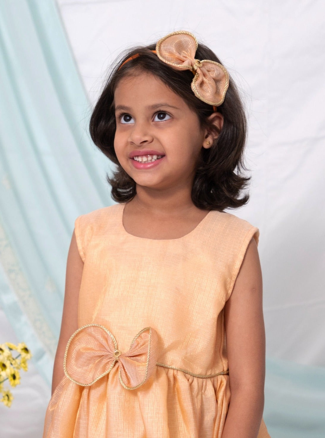 Peach tissue kotta silk sleeveless flared dress with statement embellished bow for Girl.Peach tissue kotta fabric has a subtle shimmer all over yet maintains a soft touch.The statement bow is highlighted with golden chord and waist belt has cute flowers detailing at the ends.