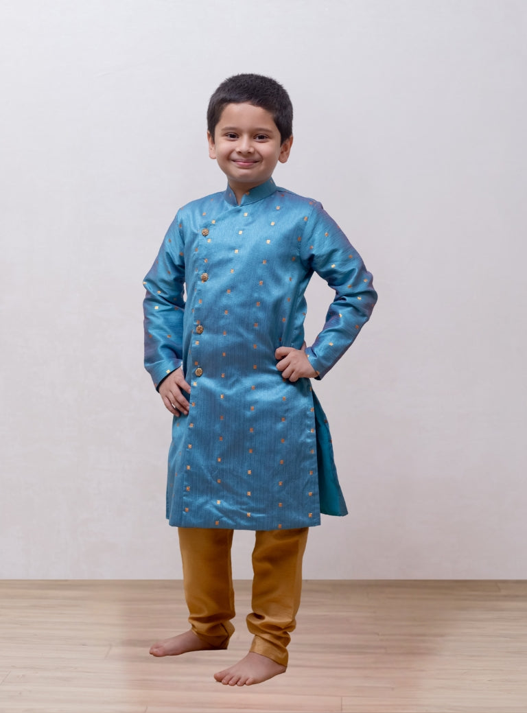 This boys' kurta features a Steel Blue mulberry silk fabric with a front open stand collar and distinctive stone-studded buttons. It is paired with a Golden Banglori silk salwar, and the kurta includes a comfortable cotton lining for added comfort.
