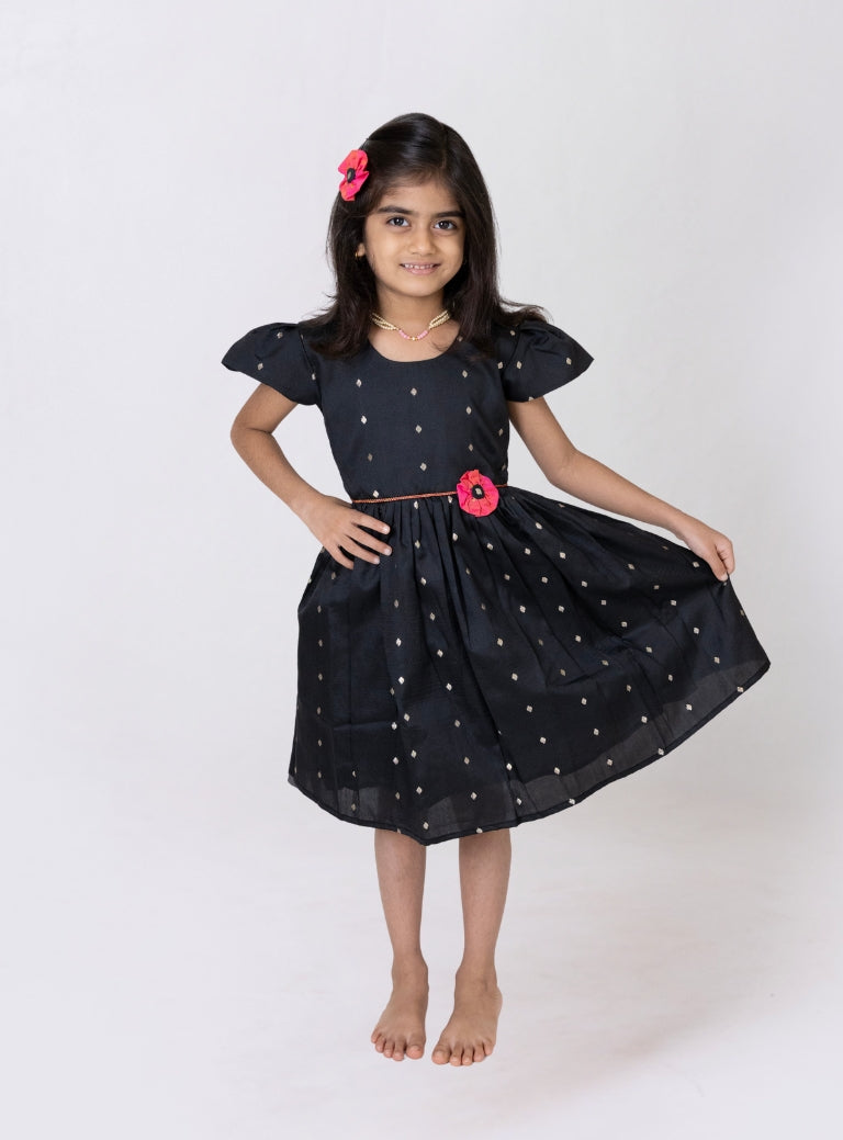 This girls’ flared dress features vibrant black taffeta silk, adorned with a handcrafted pink fabric flower motif and a chord waist belt. It is designed with elegant puffed cap sleeves for a refined finish.