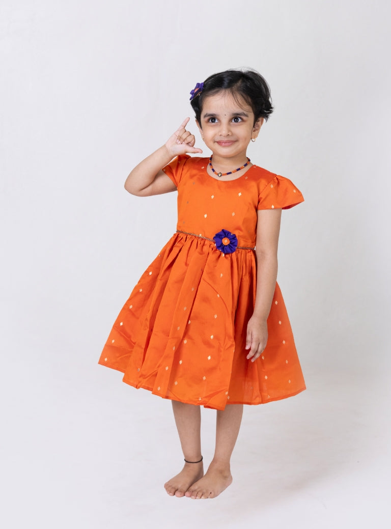 Orange taffeta silk flared dress with handmade purple fabric flower motif and chord waist belt for Girls.Let your princess be as comfortable as in her casuals with carefully designed and crafted Comfort Ethnic Wear by Soyara Ethnics. Keep her fashion quotient high with timeless patterns, vibrant combinations and royal textiles.