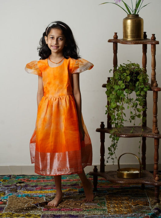 Orange organza dress with subtle shibori print and woven silver jari border for Girls.Let your princess be as comfortable as in her casuals with carefully designed & crafted Comfort Ethnic Wear by Soyara Ethnics.Keep her fashion quotient high with timeless patterns, vibrant combinations and royal textiles.